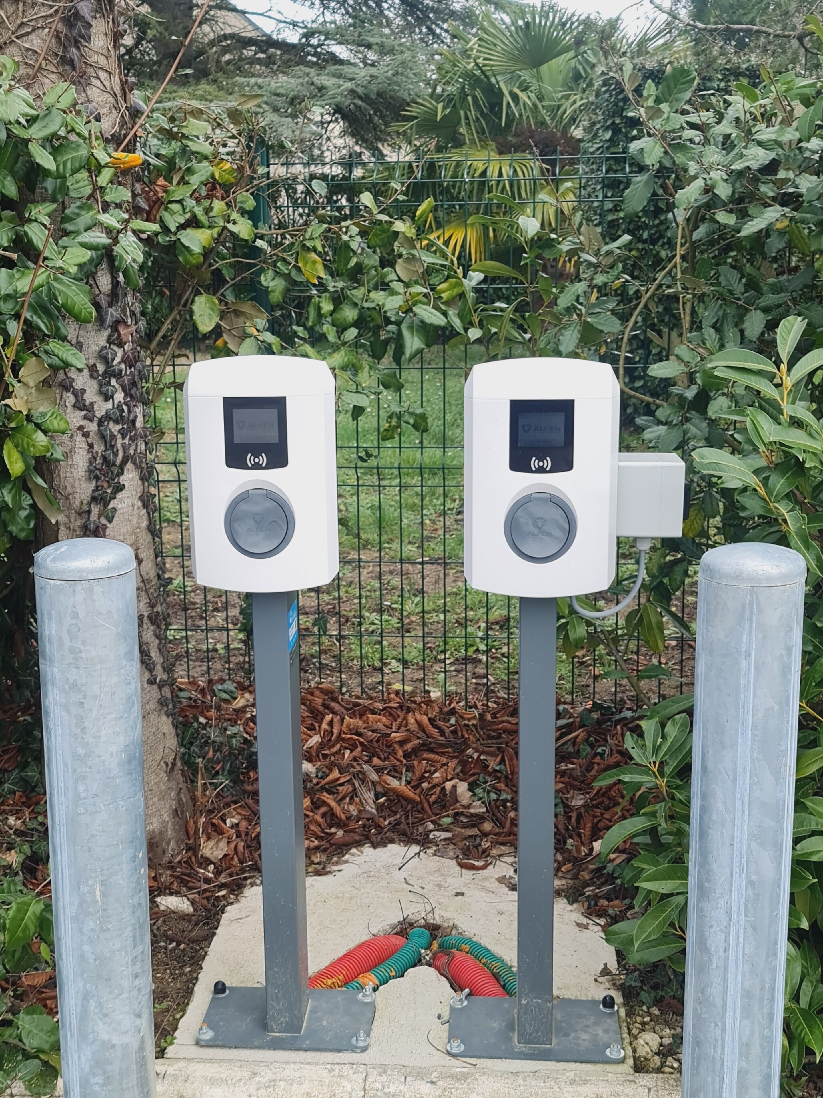 Zwei elektronische Mauterfassungssysteme an Metallpfählen, im Hintergrund ein grüner Zaun und Grünpflanzen.
