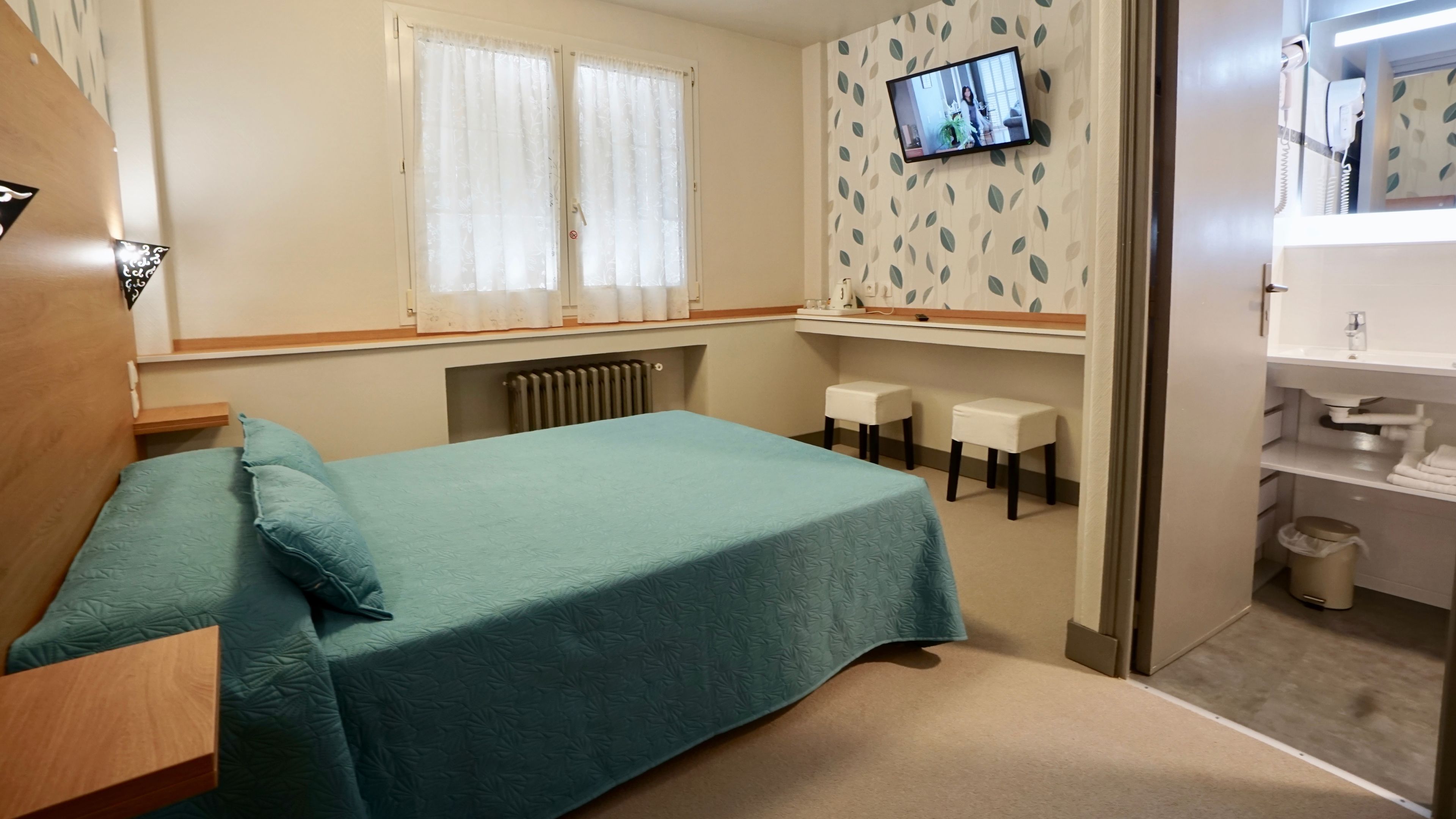 Una habitación de hotel con cama, televisión montada en la pared, radiador y una pequeña zona de estar con taburetes.