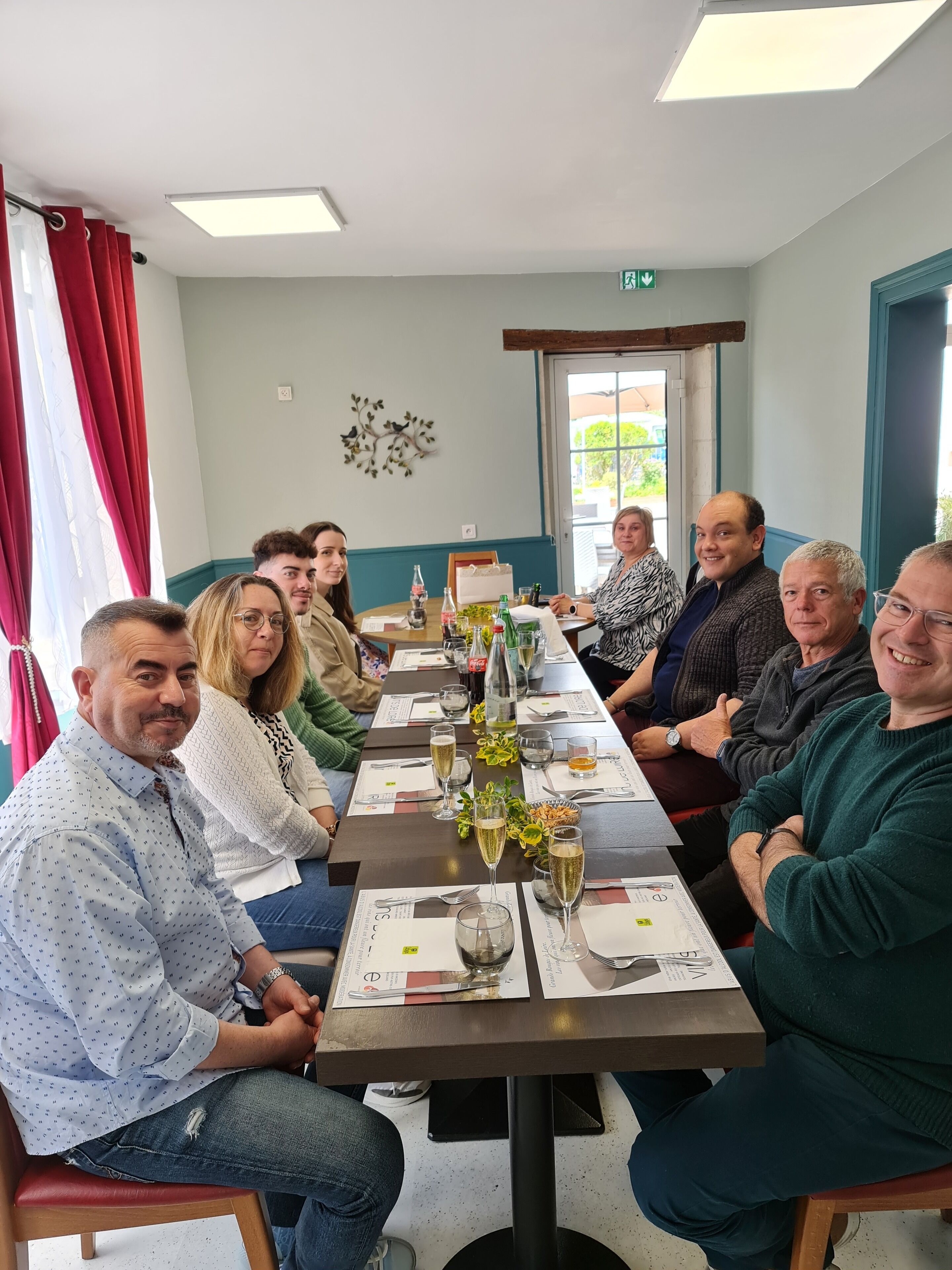 A group of people seated around a long dining table in a room with a window and a wall-mounted decoration.