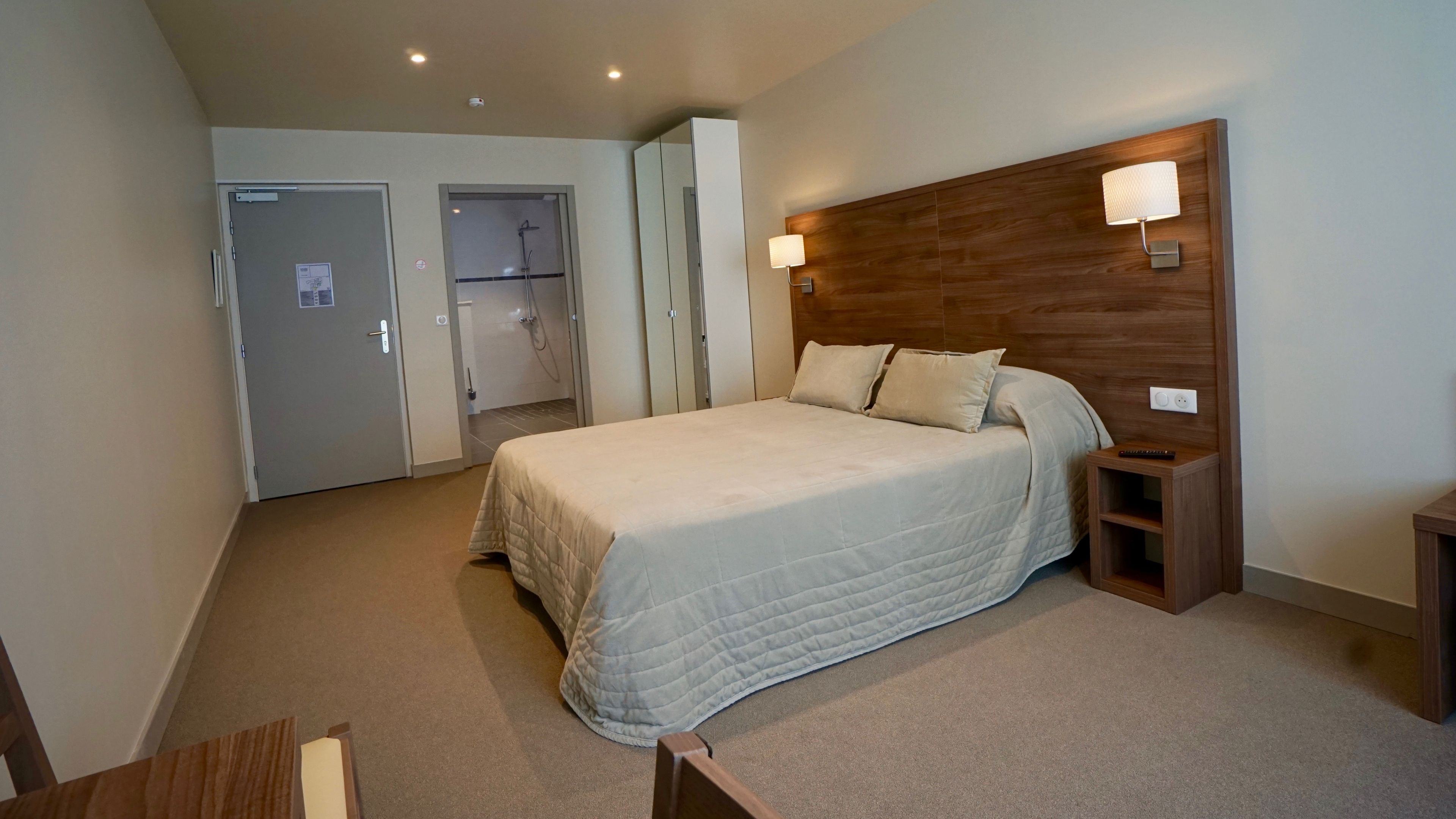 Une chambre d’hôtel avec un lit, une table de chevet et une tête de lit en bois. On y trouve une porte donnant accès à une salle de bain et un porte-manteau.