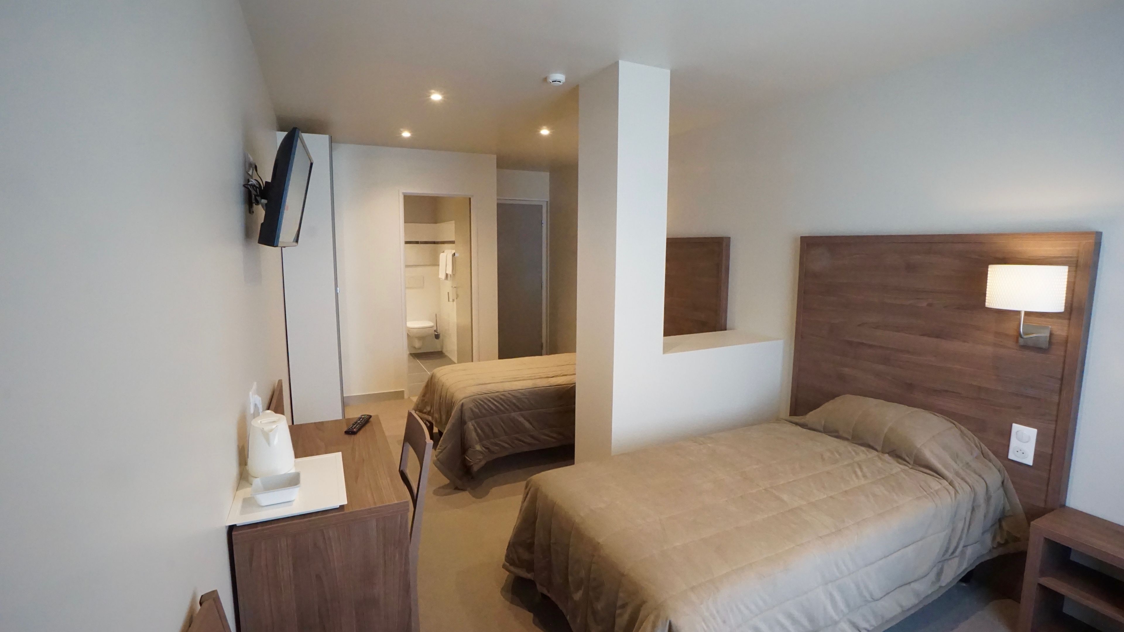 A hotel room with a bed, a desk, a chair, a TV mounted on the wall, and a bathroom visible through an open door.