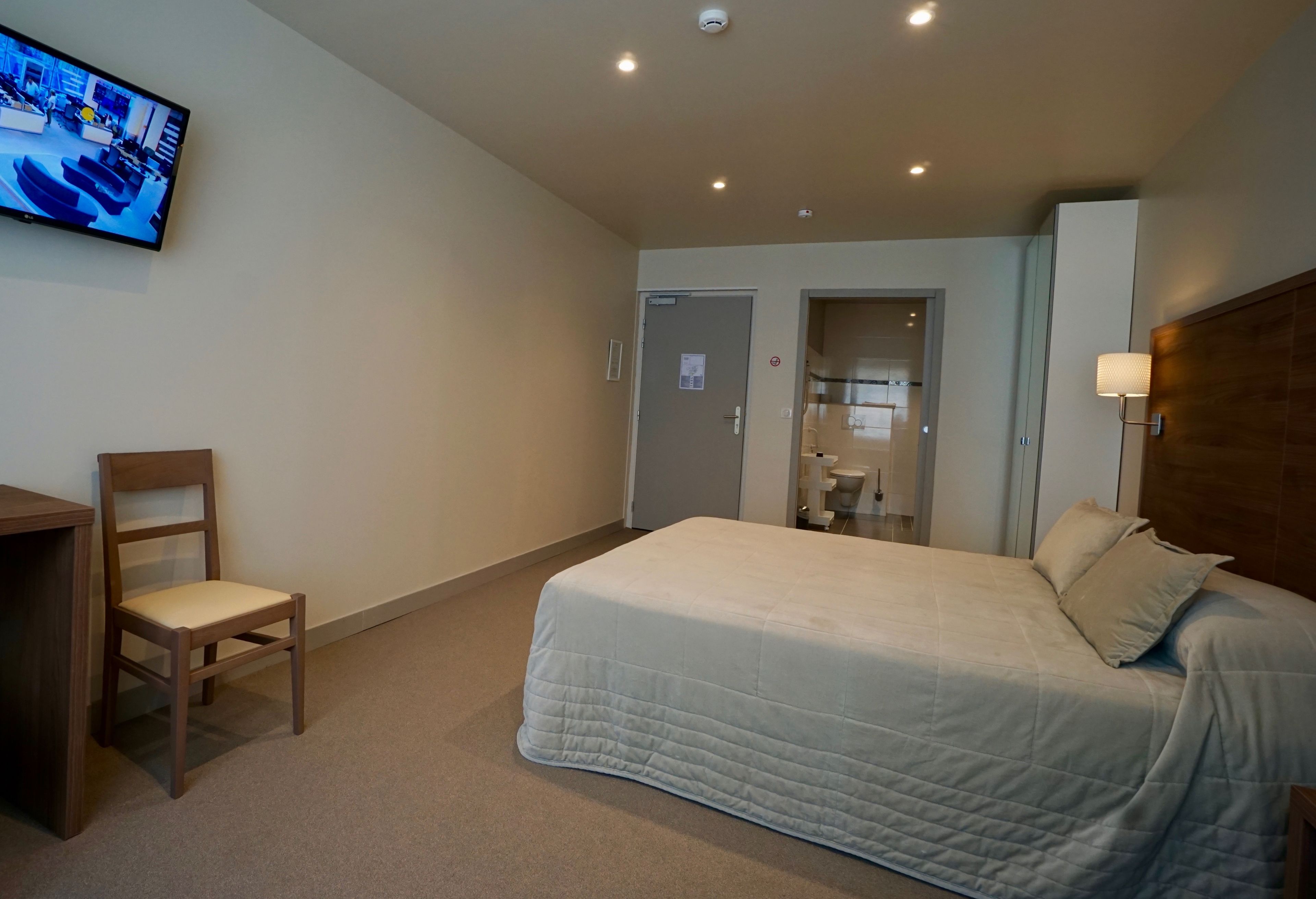 Ett hotellrum med en säng, en stol, ett skrivbord, en tv och ett badrum som syns genom en öppen dörr.