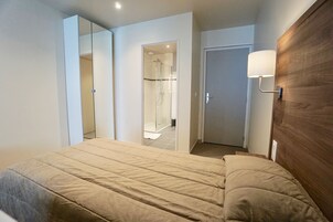 Une chambre à coucher comprenant un lit, une tête de lit en bois, un miroir, une porte et un espace douche.