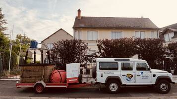 Un vehículo utilitario blanco con un remolque que transporta una cesta de mimbre y un objeto cilíndrico rojo, estacionado frente a un edificio residencial con techo de tejas y una chimenea.