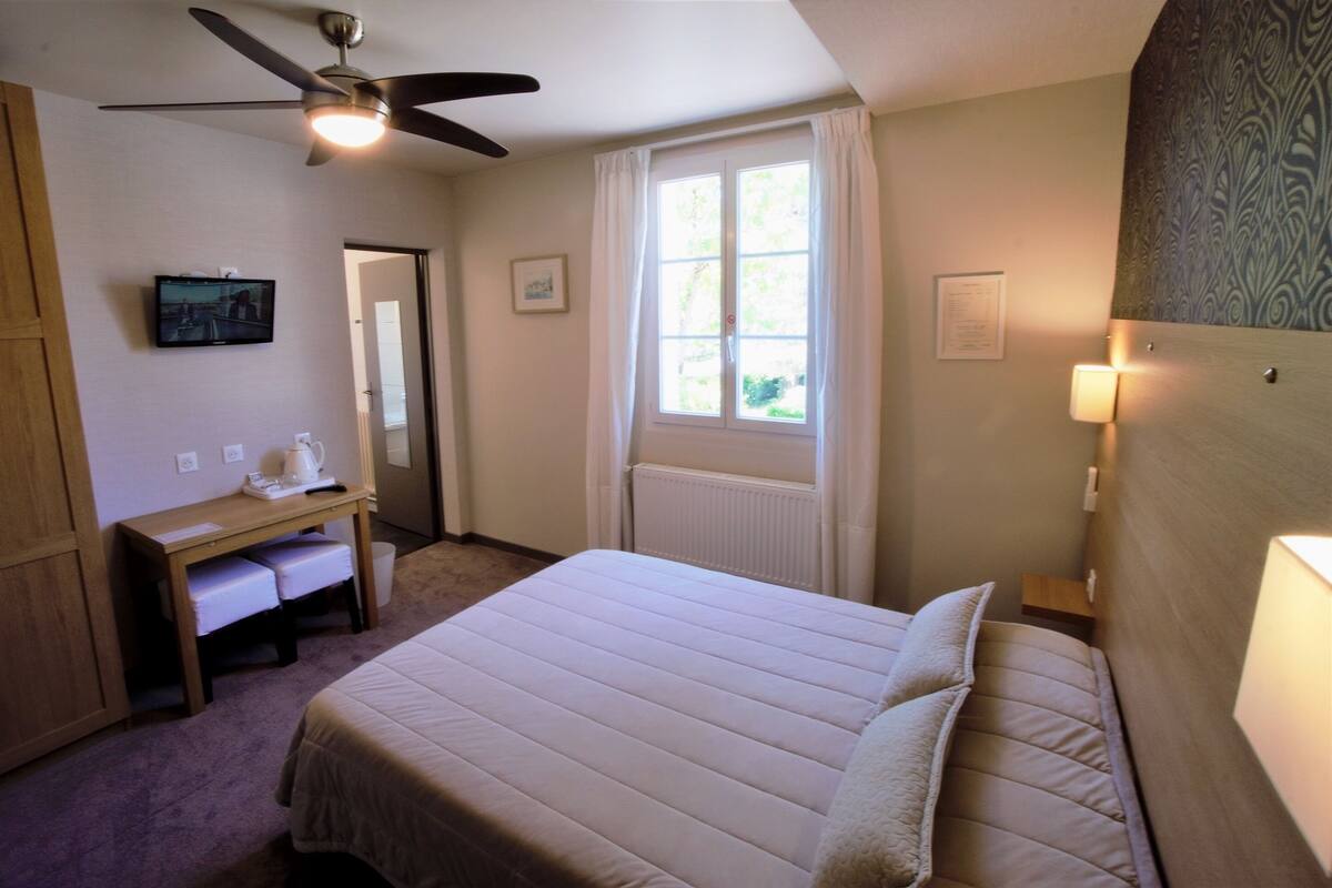 A bedroom with a bed, a desk, a ceiling fan, a window with curtains, and a wall-mounted lamp.