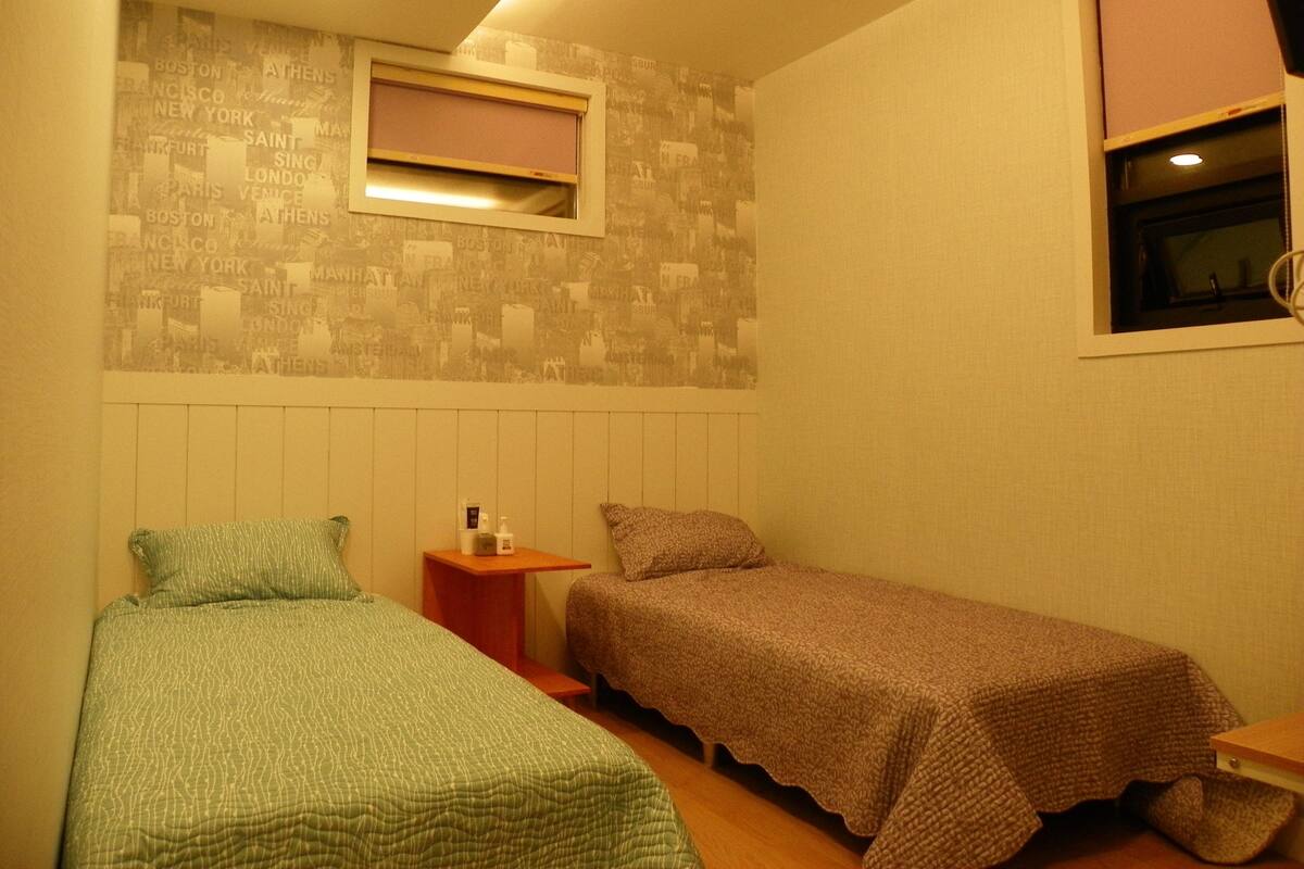 Una habitación pequeña y acogedora con dos camas individuales, una mesita de noche, un estante en la pared y una ventana con persiana.