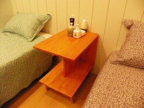 Ein kleines Zimmer mit zwei Betten, einem Nachttisch aus Holz mit Pflegeartikeln und einem gemusterten Kissen.