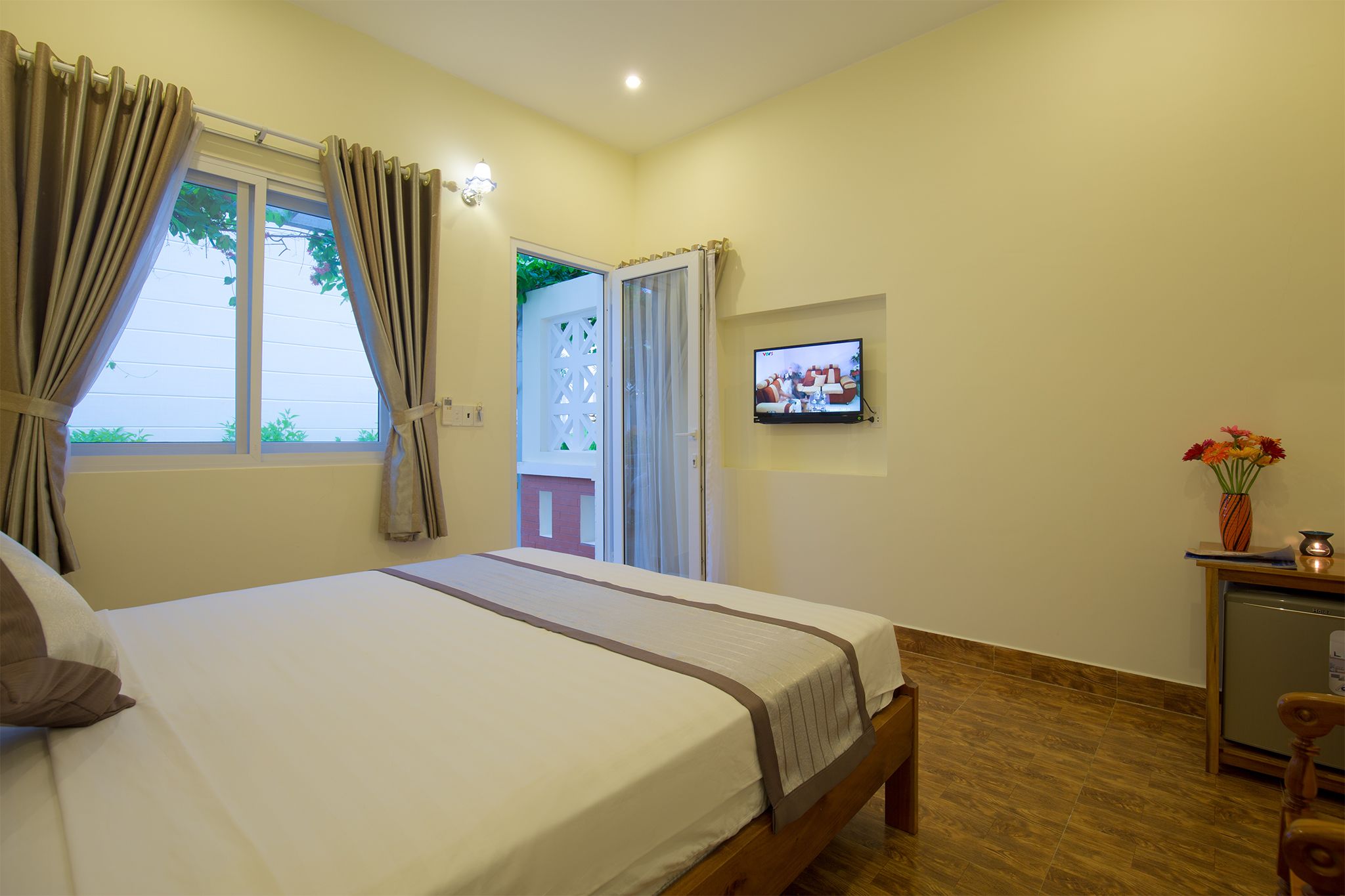 A hotel room with a bed, curtains, a television, a small table with a vase, and a door leading outside.