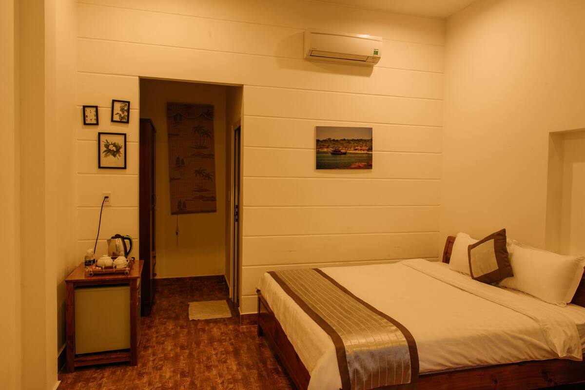 A hotel room with a bed, a small table with a kettle, a picture on the wall, and an air conditioner.