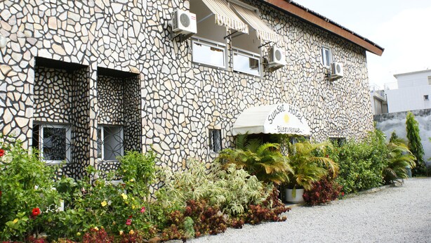 A stone building with multiple air conditioning units, a sign reading "Sunset Hotel," and a variety of potted plants and flowers in the foreground.