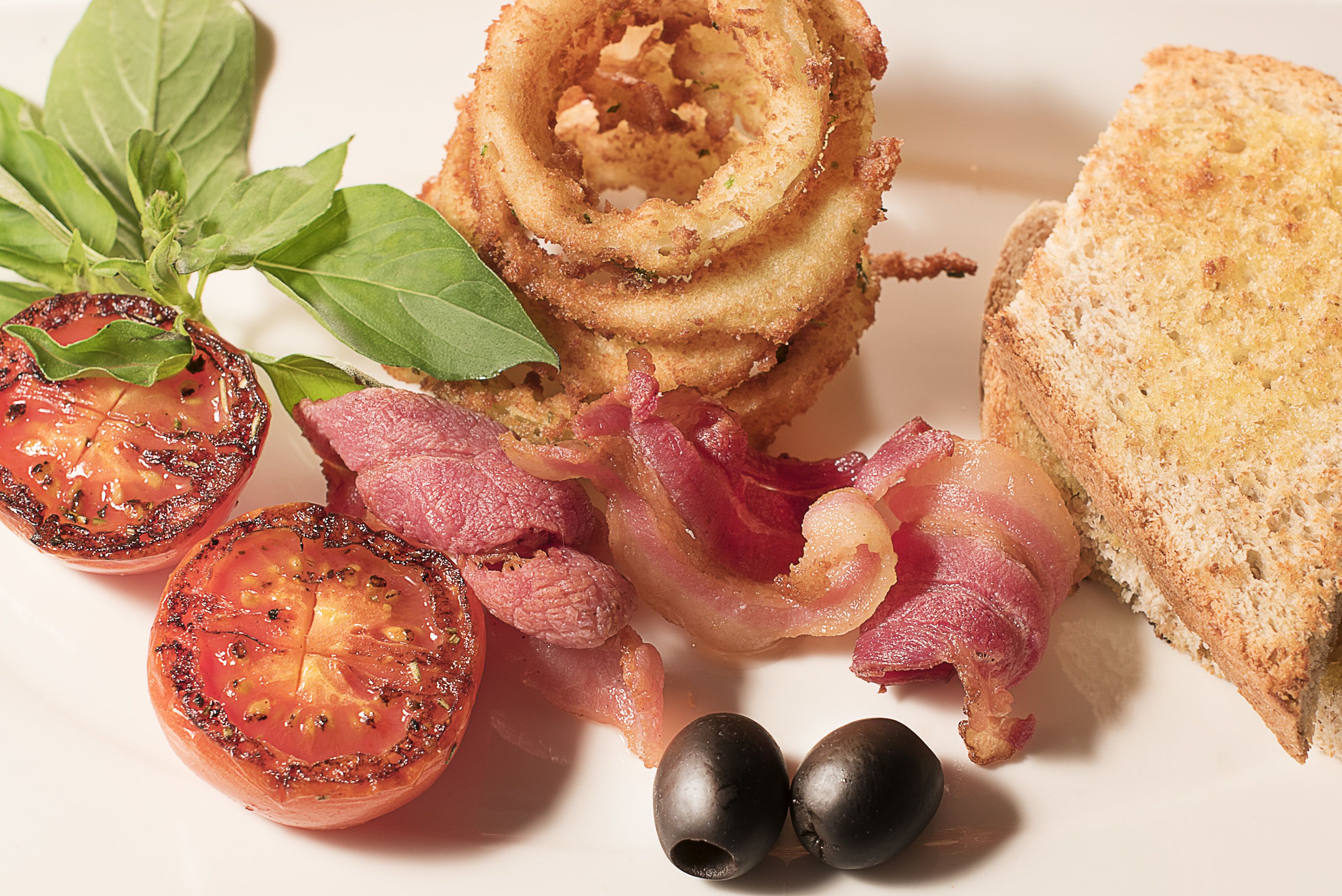 A plate with grilled tomatoes, bacon, black olives, and bread.