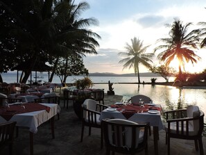 An outdoor dining area with tables set for two, overlooking a body of water with palm trees and a sunset in the background. - Gangga Island Resort & Spa (Pulau Gangga)