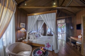 A spacious bedroom with a four-poster bed, a wicker chair, a bedside table with a lamp, and a view of the outdoors. - Gangga Island Resort & Spa (Pulau Gangga)