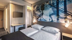 Une chambre d’hôtel moderne avec un grand lit, un téléviseur fixé au mur et une fresque murale représentant un cycliste.