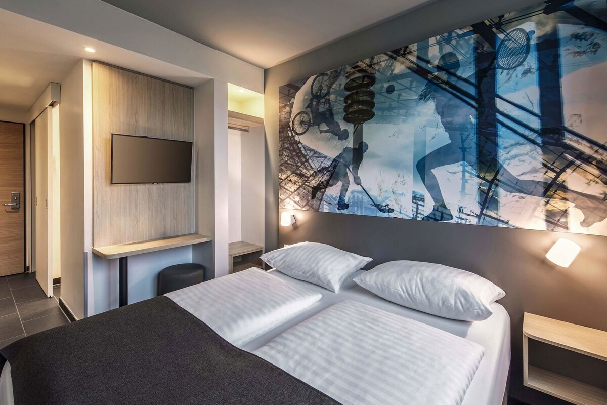 Quarto de hotel moderno com uma cama grande, televisão de tela plana na parede e uma obra de arte abstrata e original sobre a cama.
