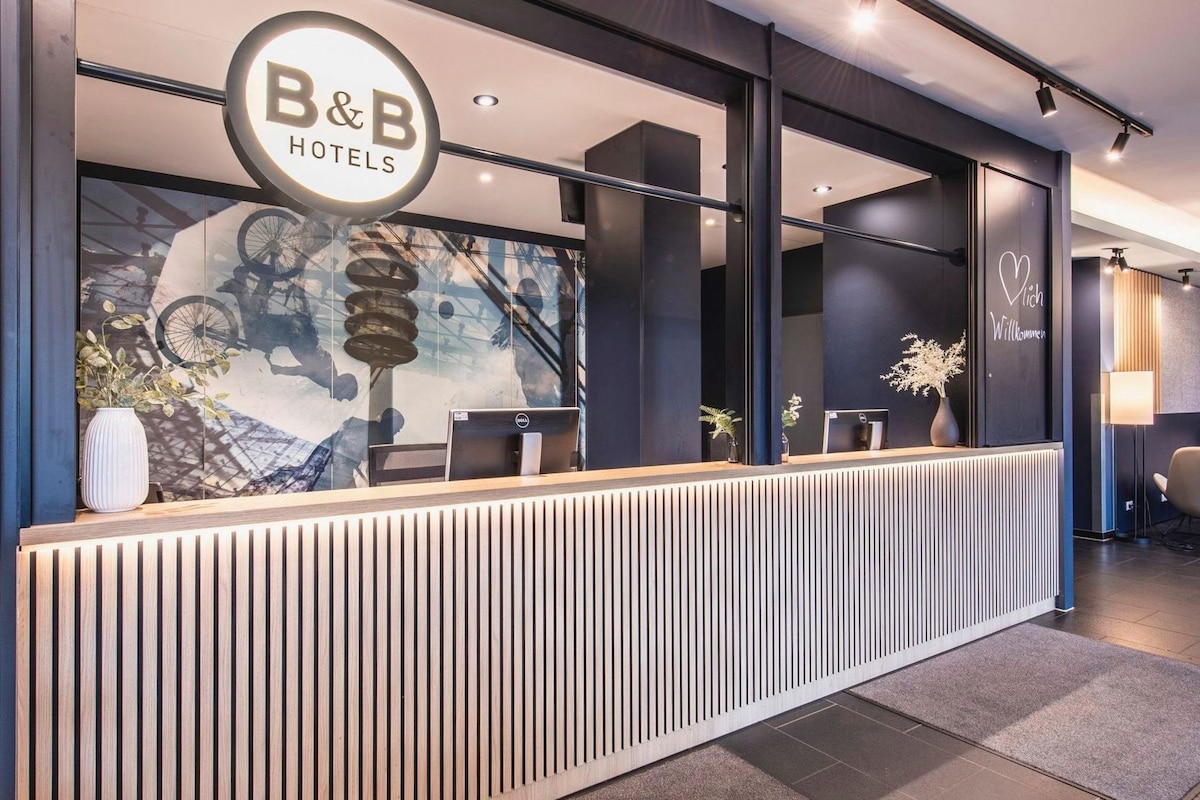 Uma receção com o logotipo B&B Hotels, um computador e uma parede decorada com um desenho de bicicleta e montanhas.