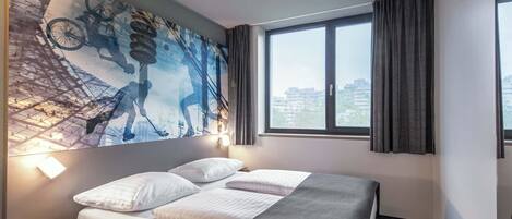 A hotel room with a bed, a large abstract painting on the wall, a window with curtains, and a view of buildings outside.