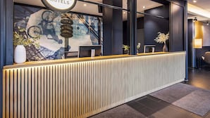 Un comptoir d’accueil orné d’une enseigne B&B Hotels, un design moderne avec des lambris en bois verticaux et une grande fenêtre décorée d’une fresque représentant des vélos.