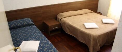 A small, compact room with two single beds, each with a blue patterned bedspread and a beige blanket. There is a wooden headboard, a small bedside table with a lamp, and a window with curtains.
