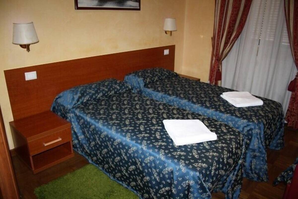 A hotel room with two single beds, a wooden nightstand, a green rug, and a window with curtains.