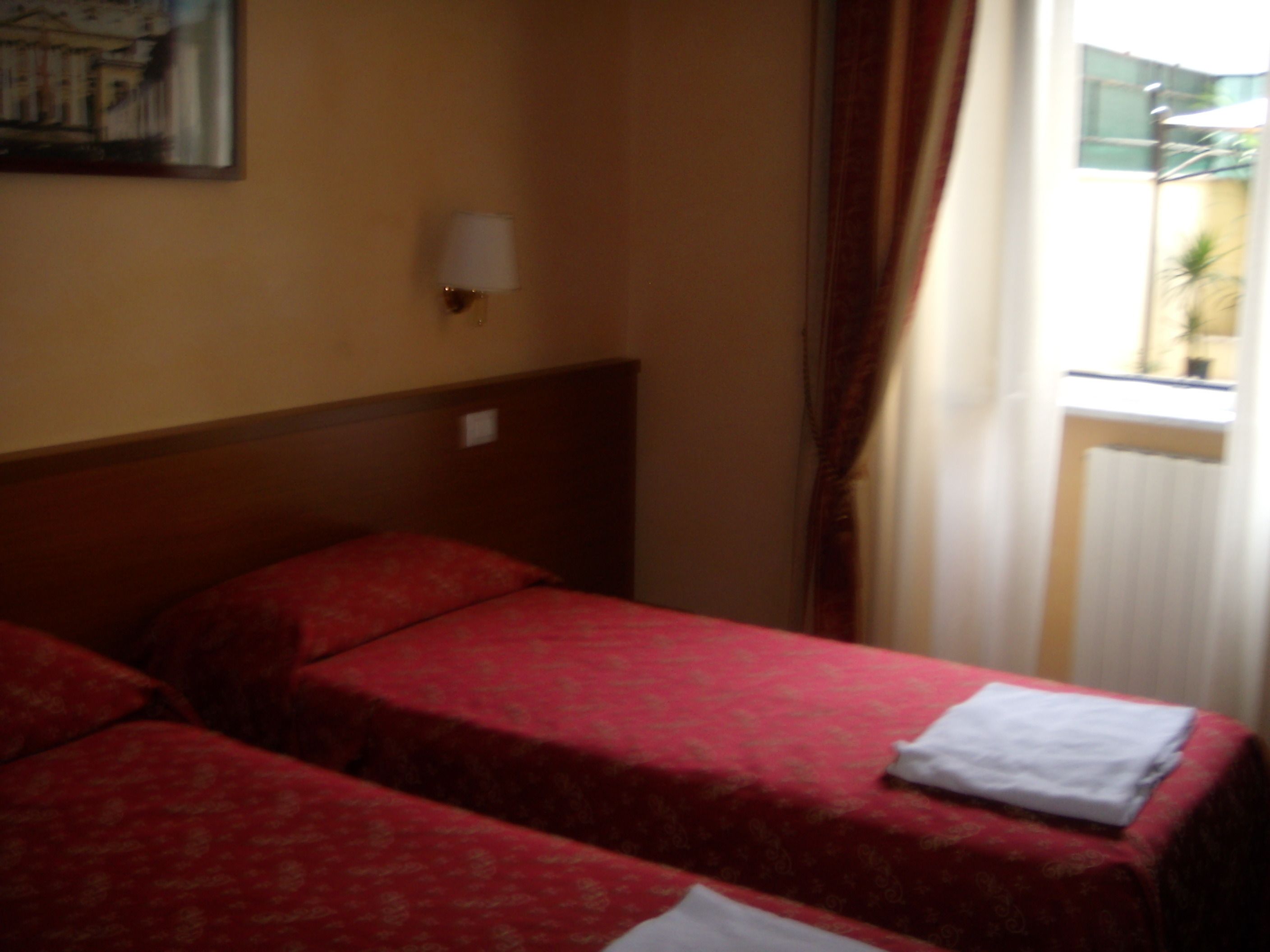 Quarto de hotel com duas camas de solteiro, mesa de cabeceira, luminária e janela com cortinas.