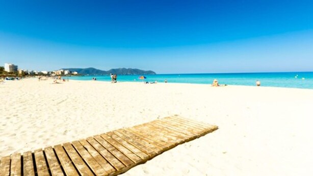 Una playa con un paseo marítimo de madera que lleva al agua, cielo azul despejado y montañas distantes.
