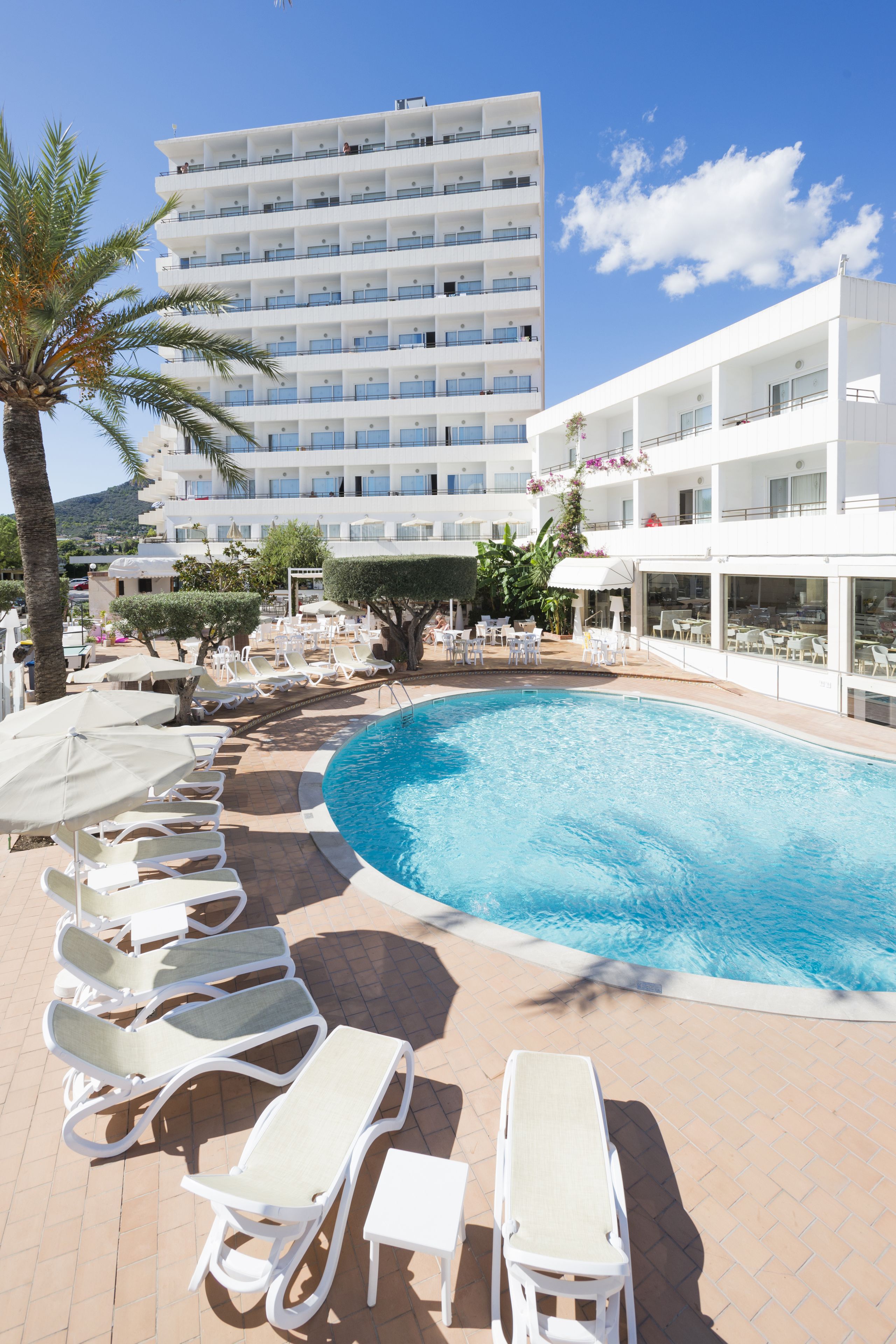 A swimming pool with sun loungers and umbrellas, surrounded by a white building with multiple balconies.