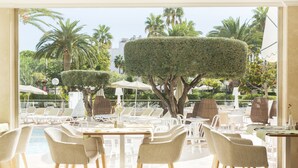 Un área de comedor al aire libre iluminada por el sol, con sillas y mesas blancas, rodeada de setos bien cuidados y palmeras.
