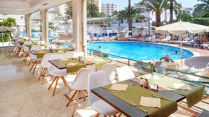 Un restaurante junto a la piscina con mesas al aire libre, una piscina y palmeras al fondo.