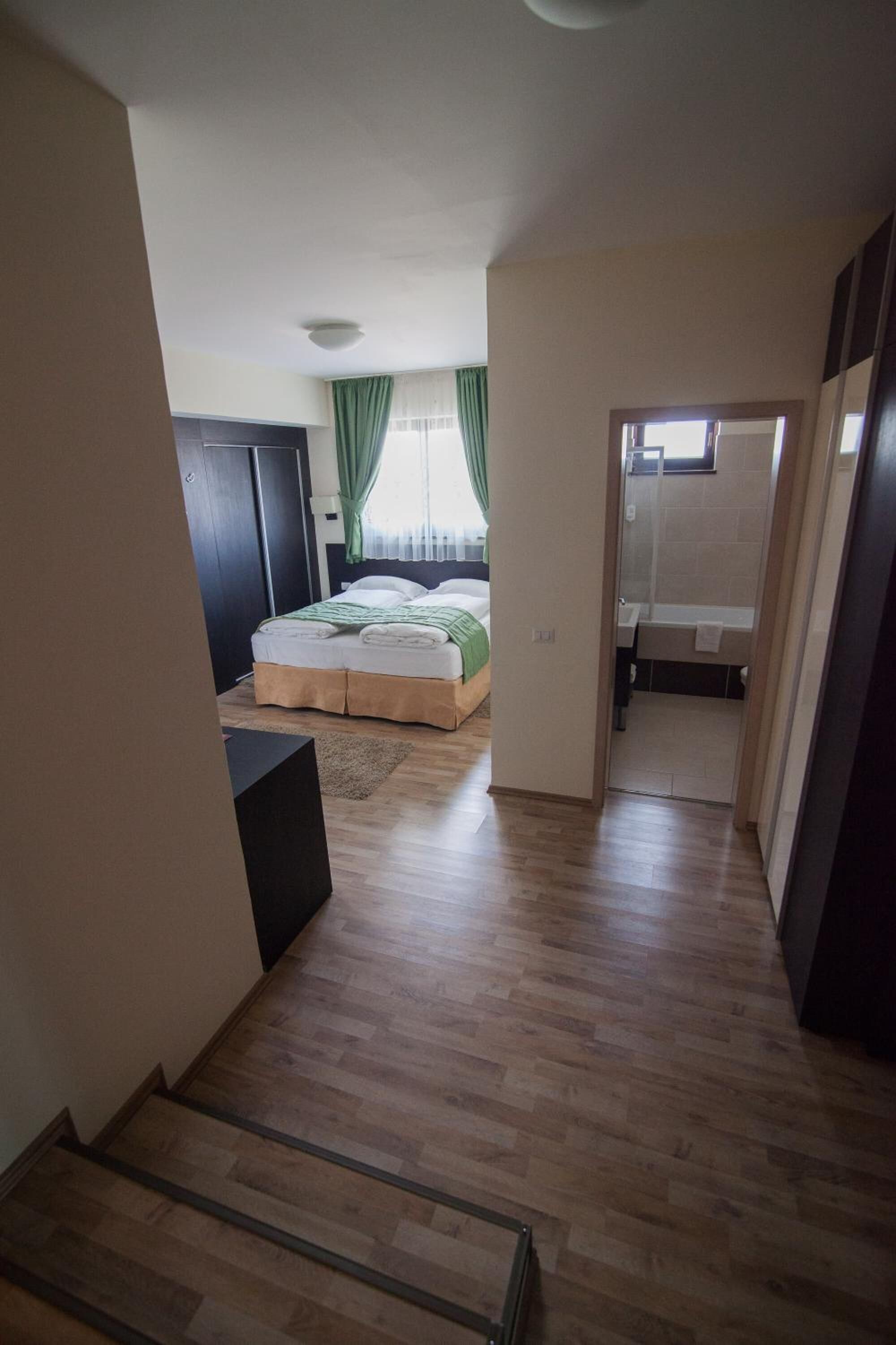 Een slaapkamer met een bed, een raam met gordijnen, een badkamer met een bad en een gang met houten vloer.