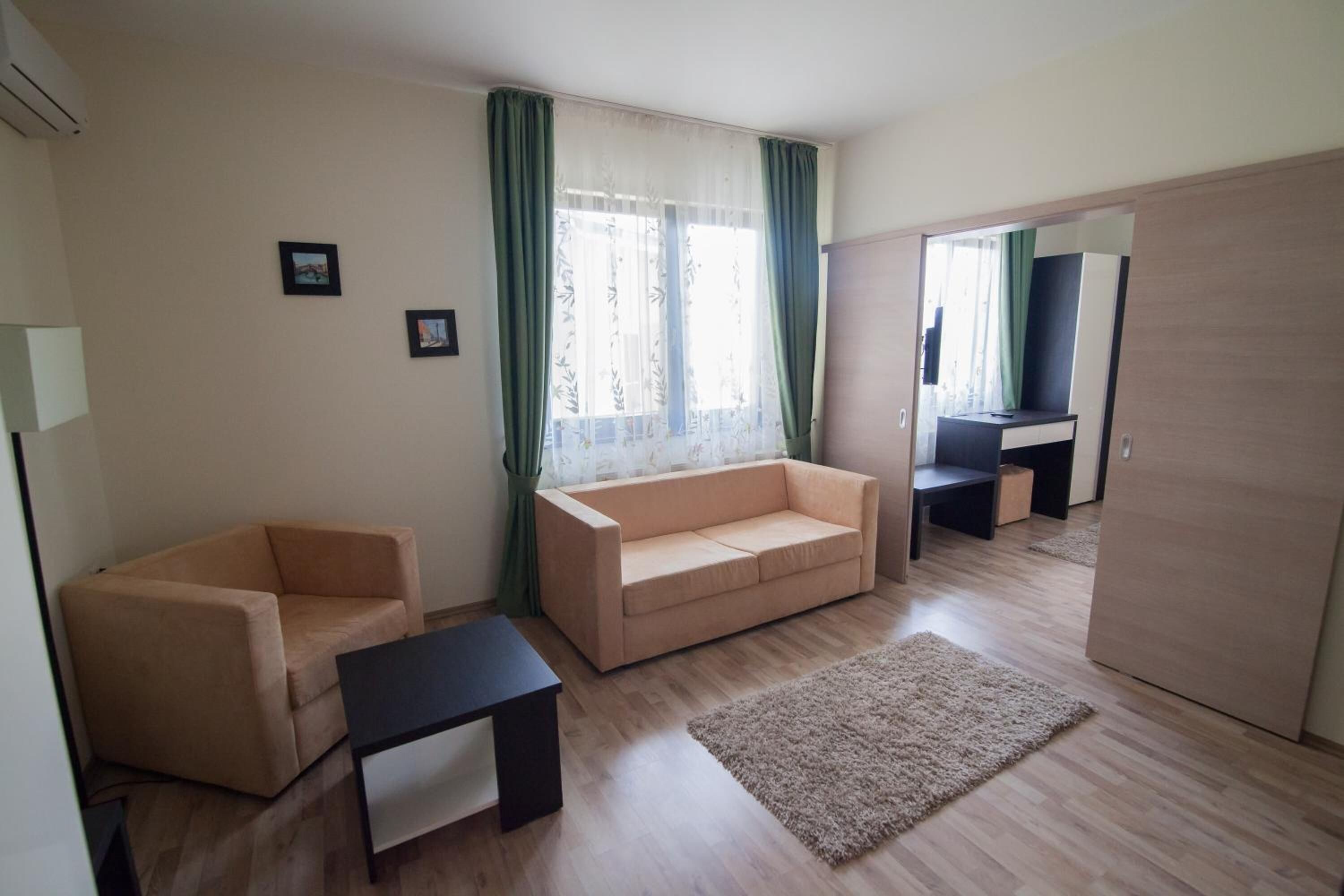 Una sala de estar con un sofá color beige, una mesita negra pequeña, piso de madera, una ventana con cortinas verdes y un televisor al fondo.