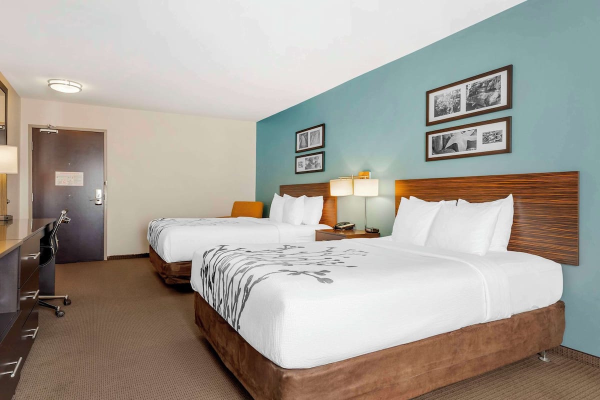 A hotel room with two beds, a desk, and a chair. There are framed pictures on the wall and a door with a sign.