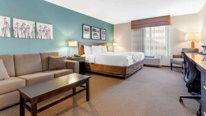 A hotel room with a bed, a sofa, a desk, and a chair. There are two lamps, a window with curtains, and artwork on the wall. - Sleep Inn & Suites Cumberland - LaVale (Cumberland)