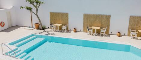 A swimming pool with steps leading into it, surrounded by white chairs and tables, and a small tree.