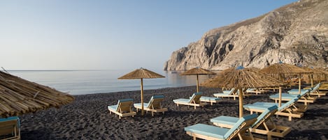 Une plage avec des chaises longues en bois et des parasols en chaume, une falaise rocheuse en arrière-plan et une mer calme.