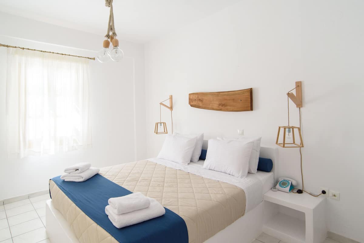 A neatly made bed with a blue and beige quilt, white pillows, and folded towels. There is a wooden headboard, a bedside table with a telephone, and a wall-mounted light fixture.