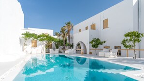A pool area with white walls and palm trees, surrounded by lounge chairs and a clear blue sky. - Santorini Crystal Blue (Santorini)