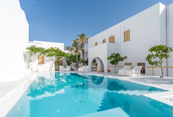A pool area with white walls and palm trees, surrounded by lounge chairs and a clear blue sky. - Santorini Crystal Blue (Santorini)