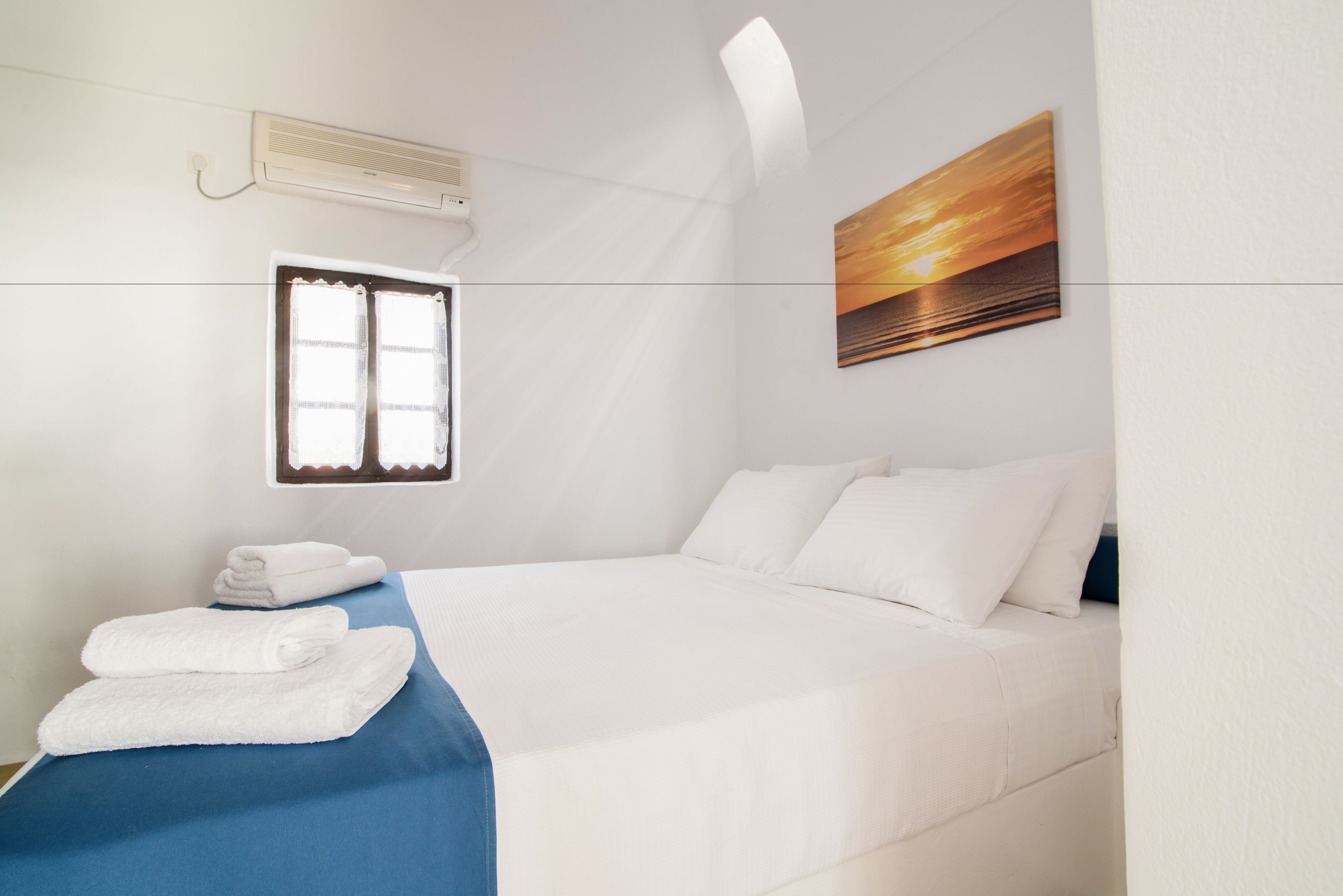 A hotel room with a bed, a window with curtains, a wall-mounted air conditioner, and a painting on the wall.