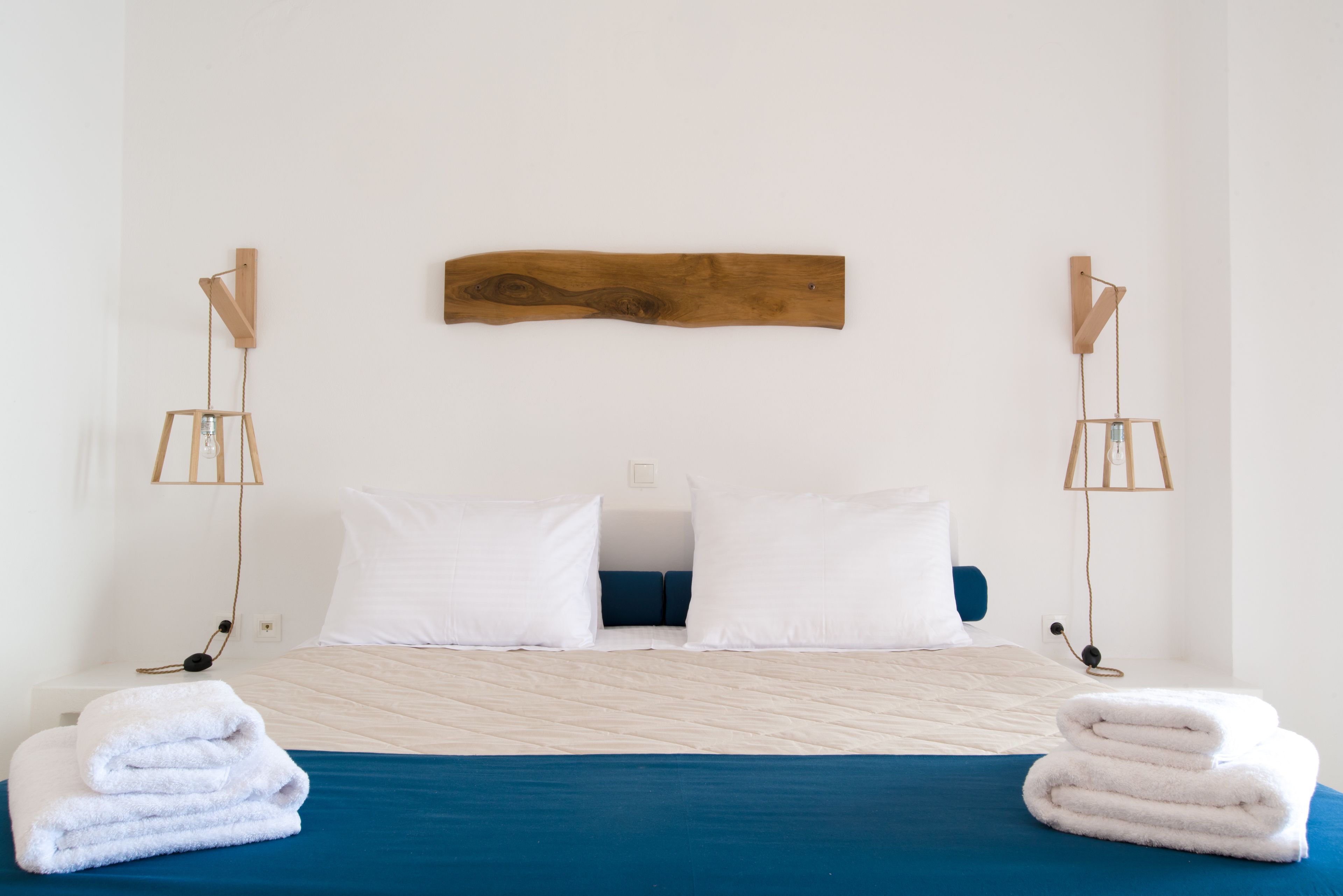 Een net opgemaakt bed met wit- en blauwe beddengoed, twee witte kussens en een houten hoofdbord. Aan beide zijden van het bed hangen twee wandlampen.