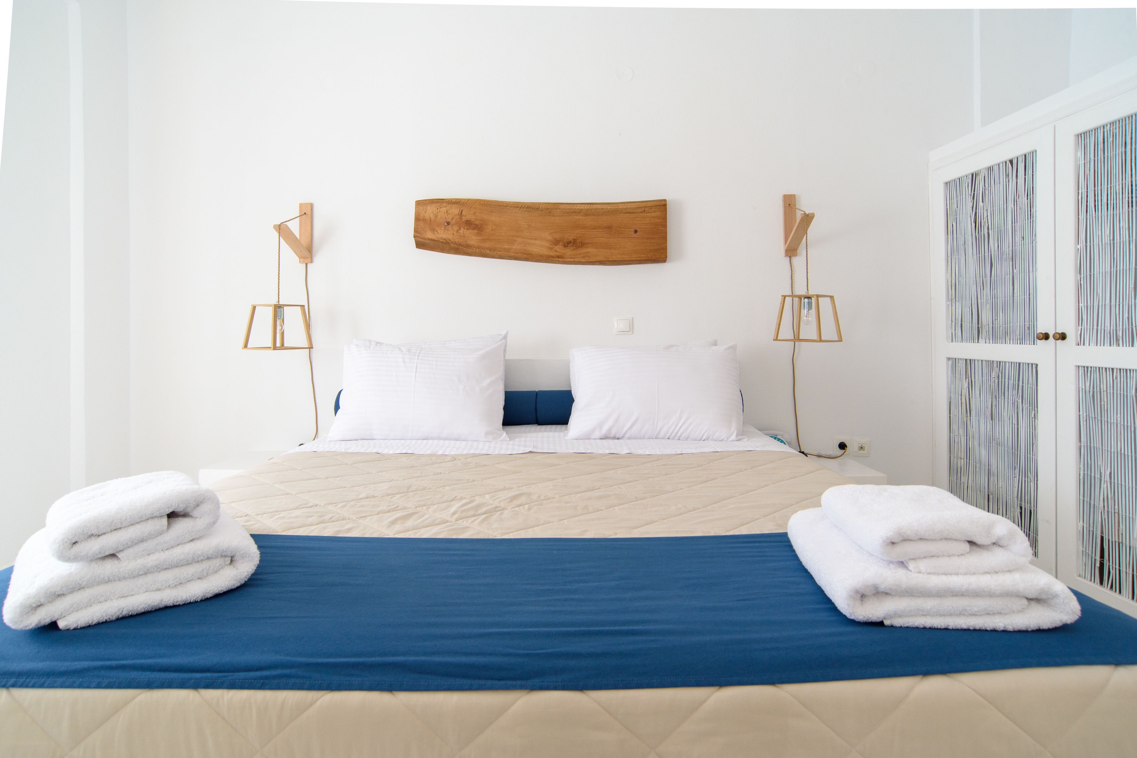 A neatly made bed with a blue bedspread and two white pillows, a wooden headboard, and a stack of white towels on the bed.