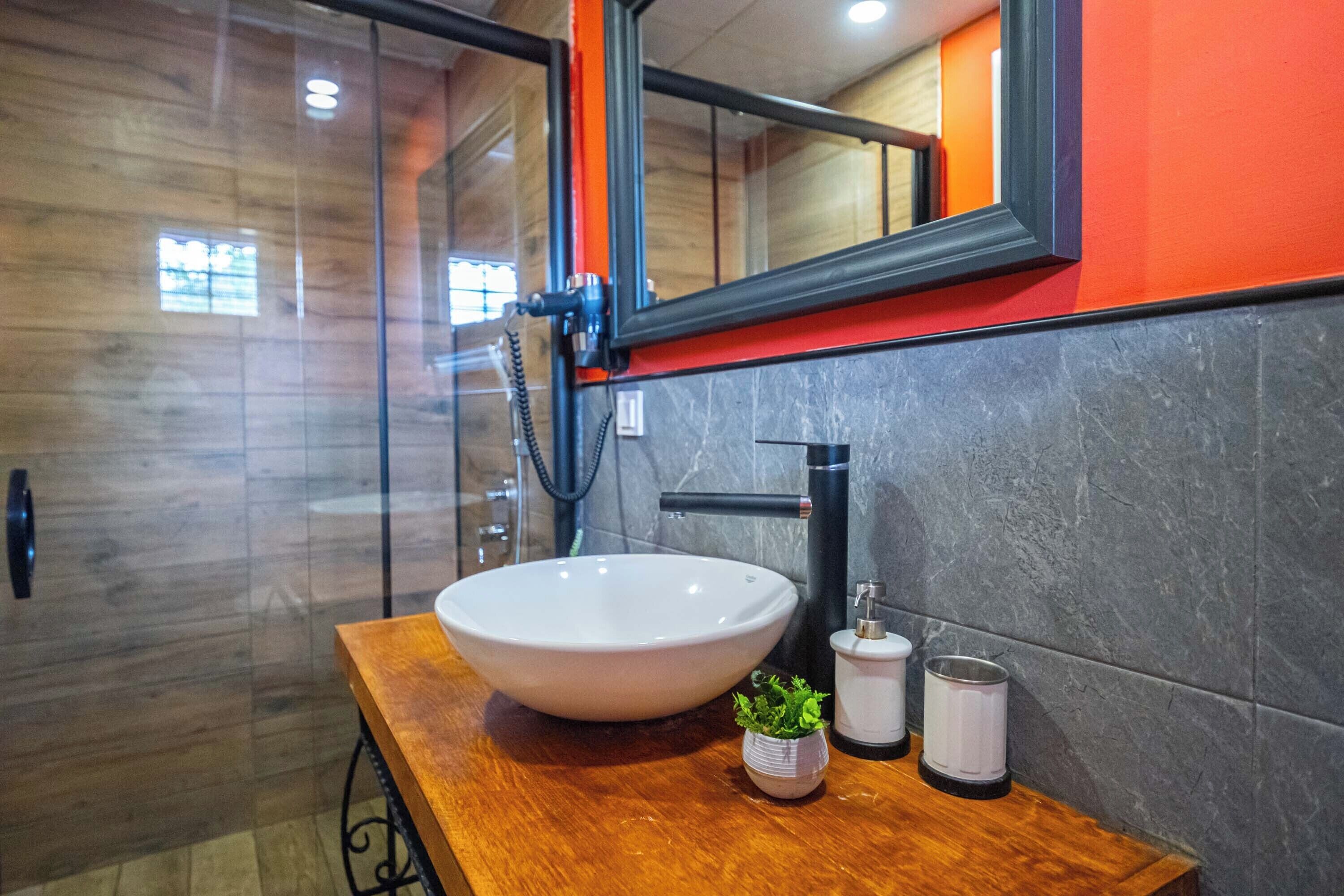A bathroom with a wooden vanity, a white vessel sink, a small potted plant, and a mirror with an orange frame.
