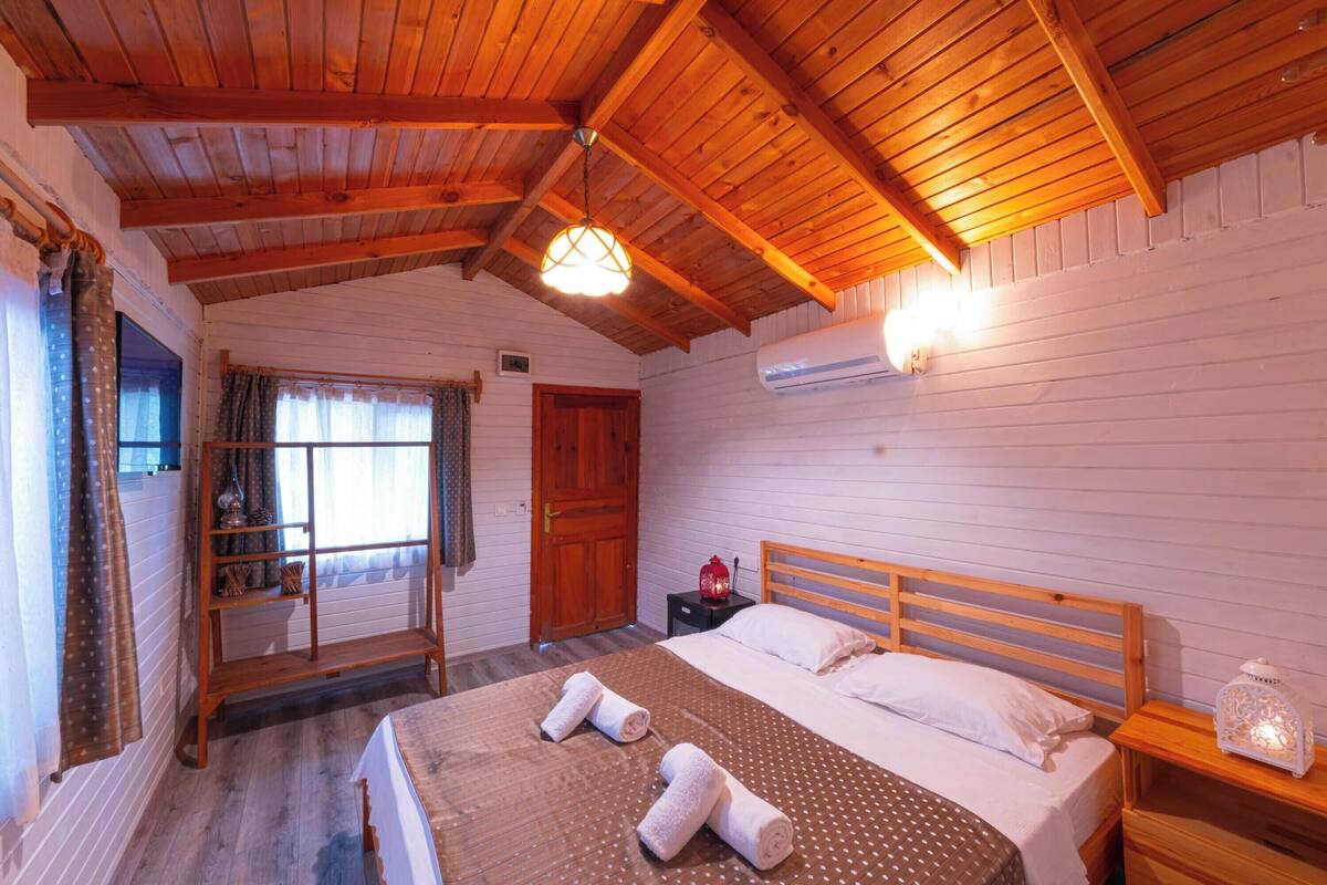 A wooden-paneled room with a bed, a nightstand, a shelf, and a window with curtains.