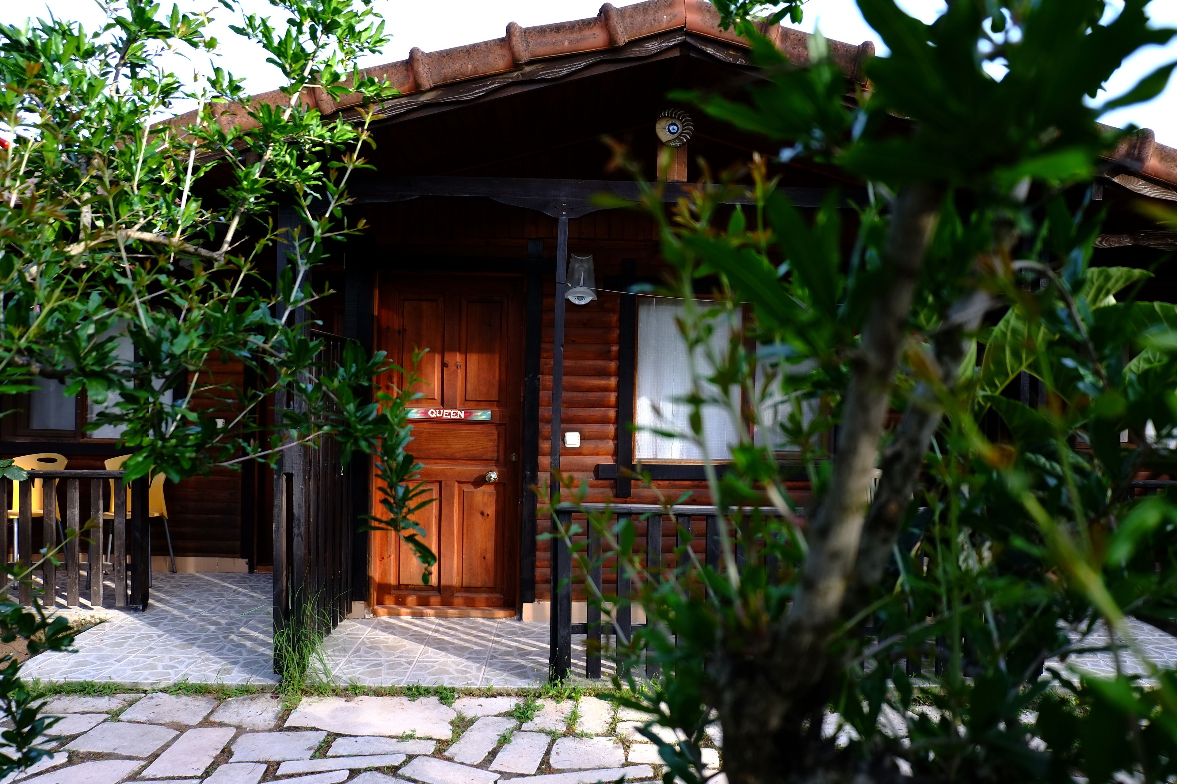 Una cabaña de madera con un acceso con baldosas de piedra, una puerta de madera con la etiqueta 'QUEEN' y una pequeña zona de estar al aire libre con sillas amarillas.