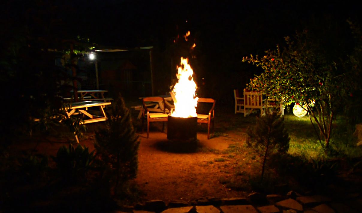 A nighttime outdoor setting with a central fire, surrounded by chairs and a table, indicating a patio or garden area.
