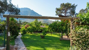 A stone pathway leading through a lush garden with a pergola overhead, surrounded by greenery and a mountain in the background. - Koala Bungalows (Kumluca)