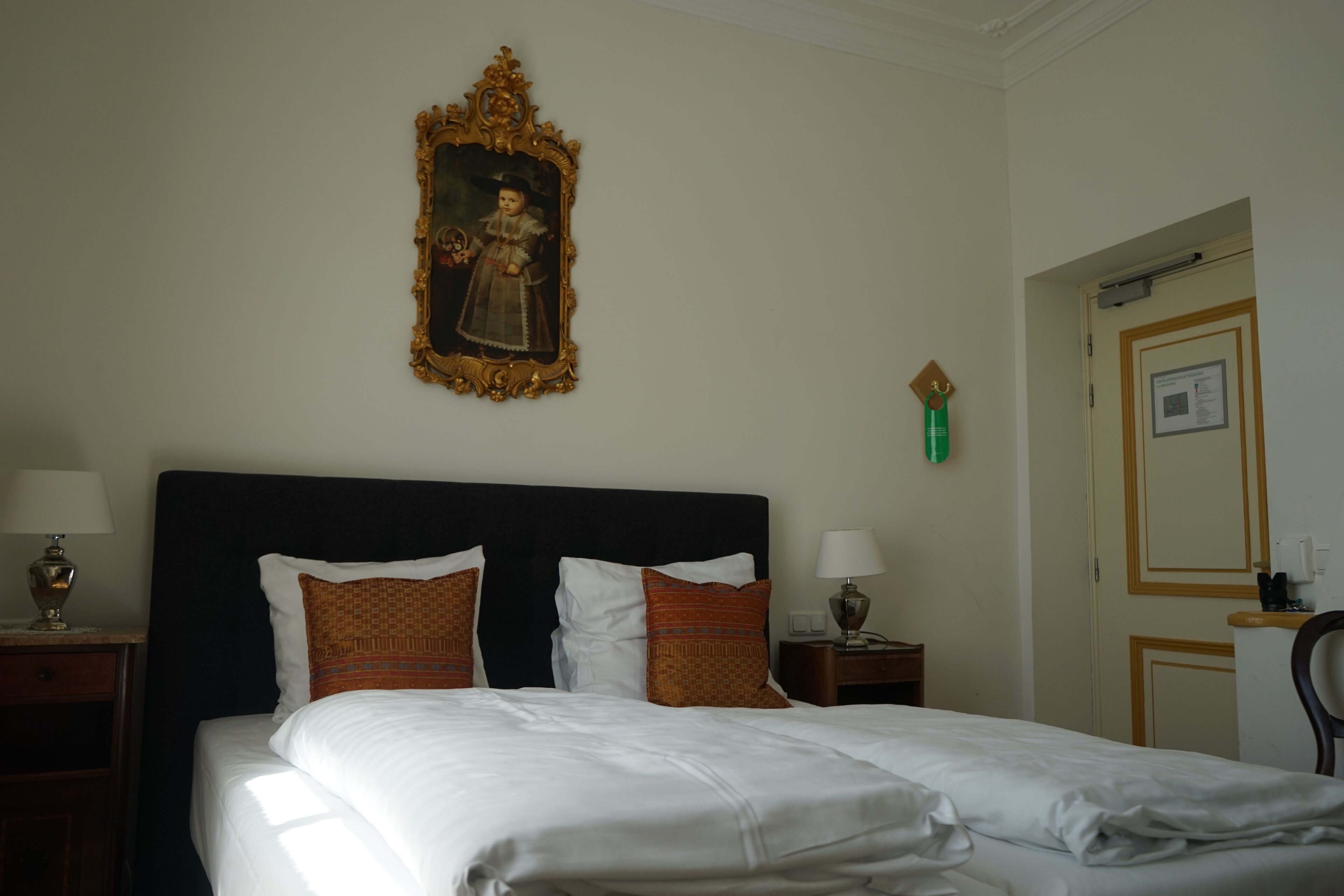 A hotel room with a large bed, two nightstands, a wall-mounted painting, a door with a mirror, and a small table with a lamp.