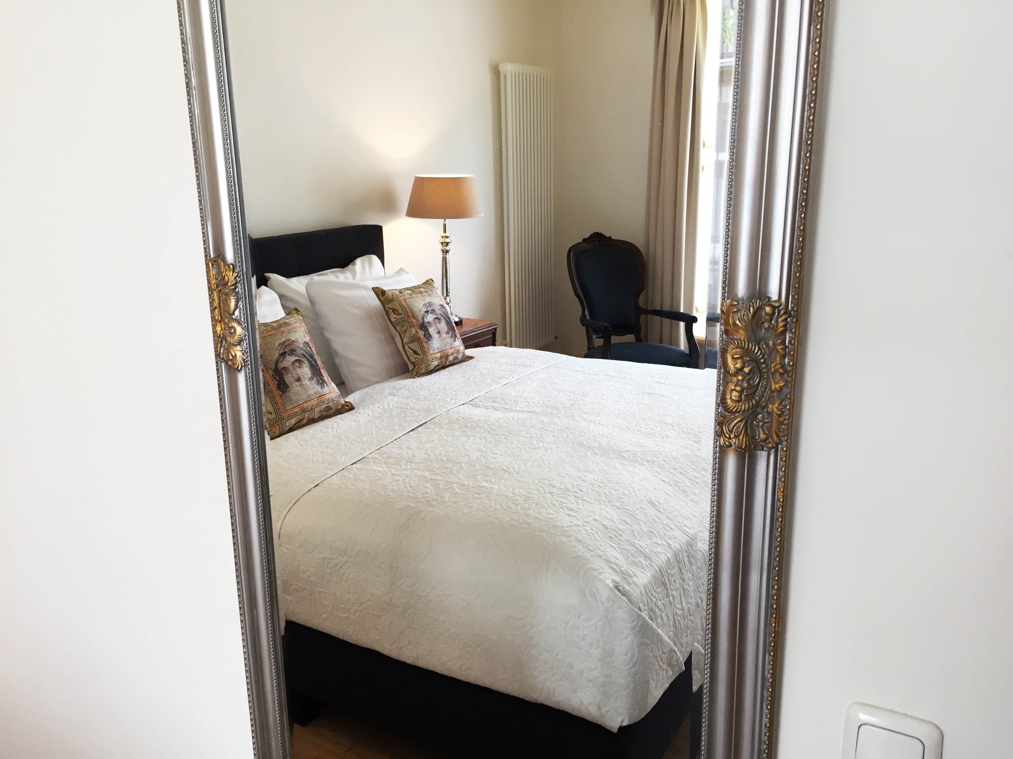 Una cama bien hecha con sábanas blancas y almohadones decorativos, una lámpara de noche y una silla en una habitación de diseño sencillo y limpio.
