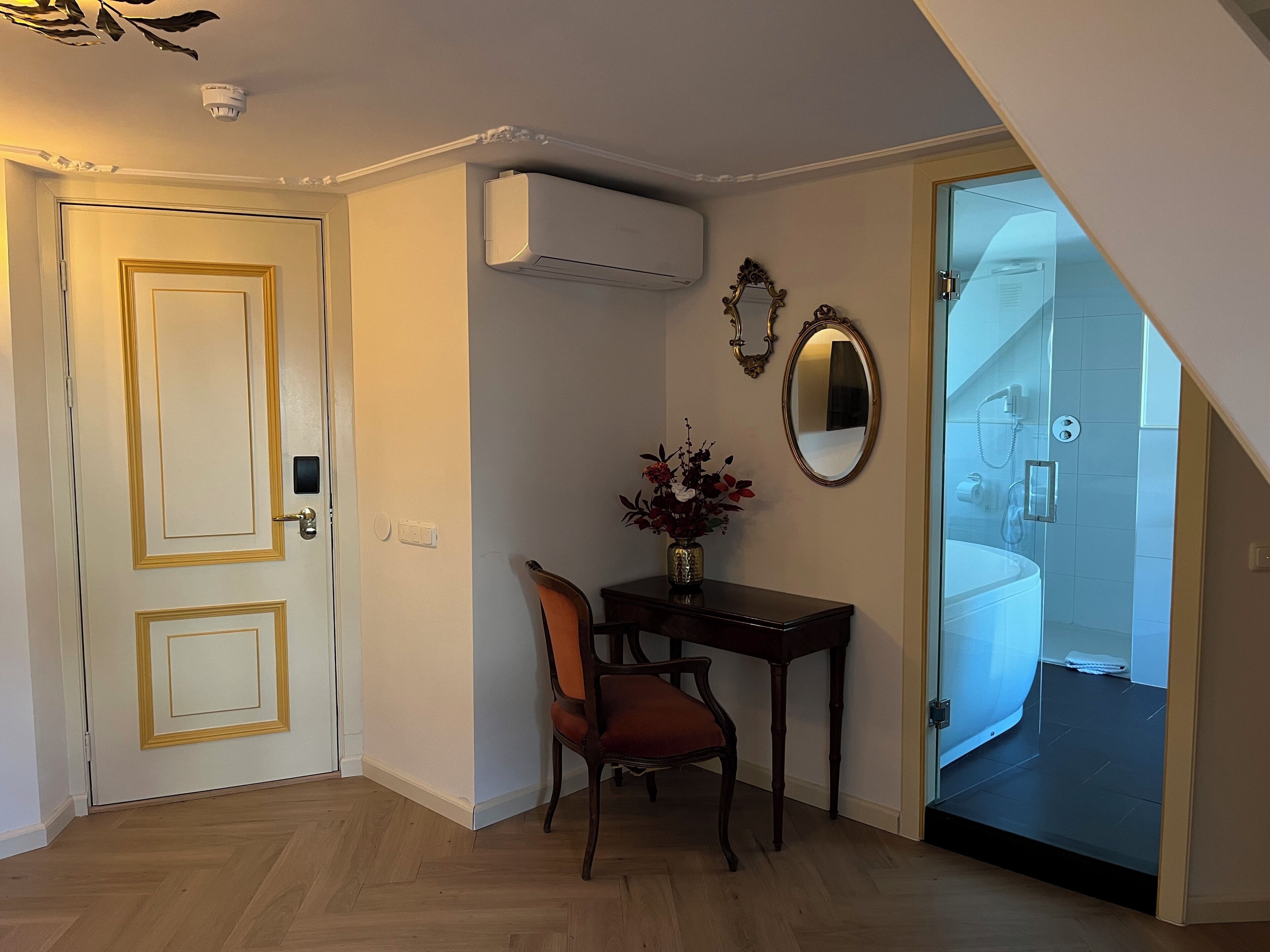 Una habitación con piso de madera, una puerta con marco dorado, una pequeña mesa con un florero de flores, una silla y un espejo en la pared.