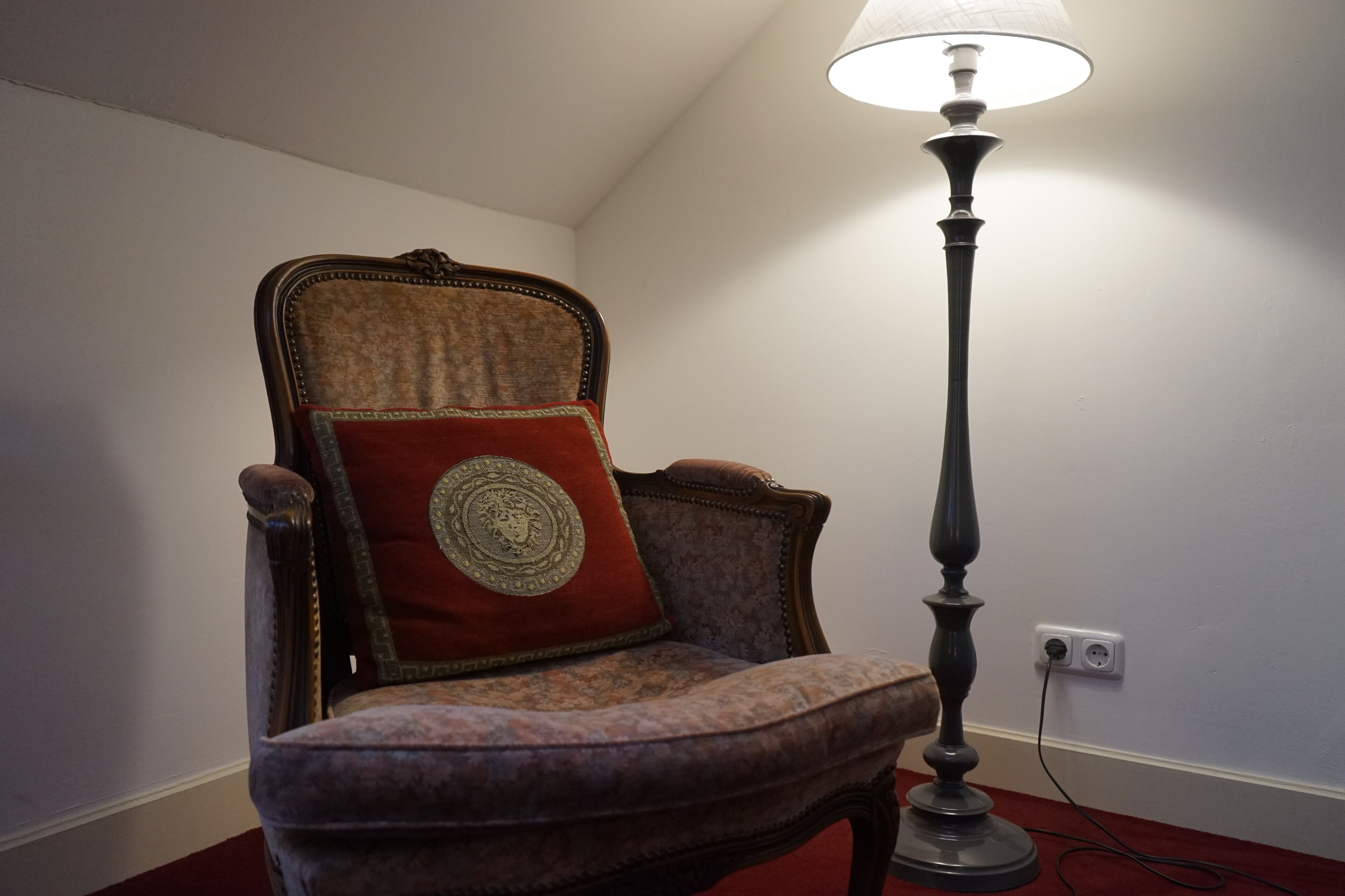 A vintage chair with a decorative cushion and a tall floor lamp in a corner of a room.